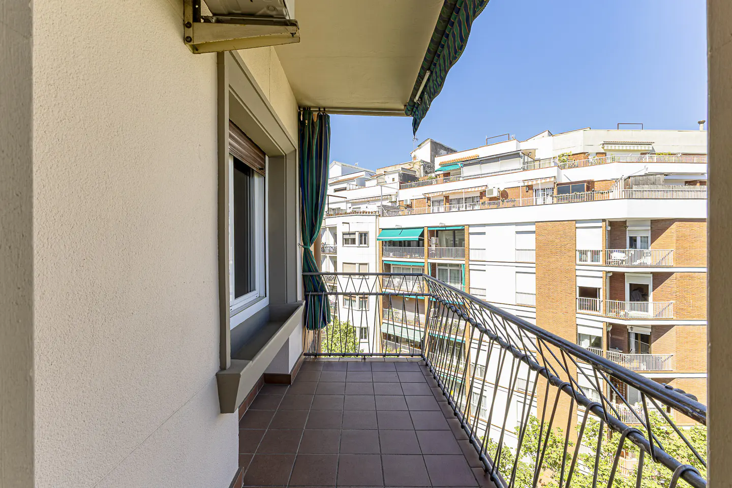 A balcony with brown tile flooring and a black metal railing overlooks city buildings under a blue sky. A window with closed blinds is on the left.