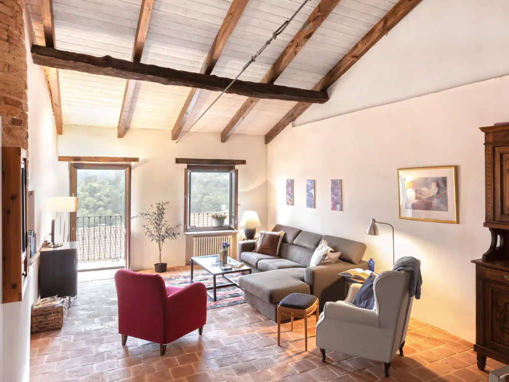 Living room with exposed beams, brick floors, and white walls. A gray sectional sofa, red chair, and white armchair furnish the space. Balcony view.