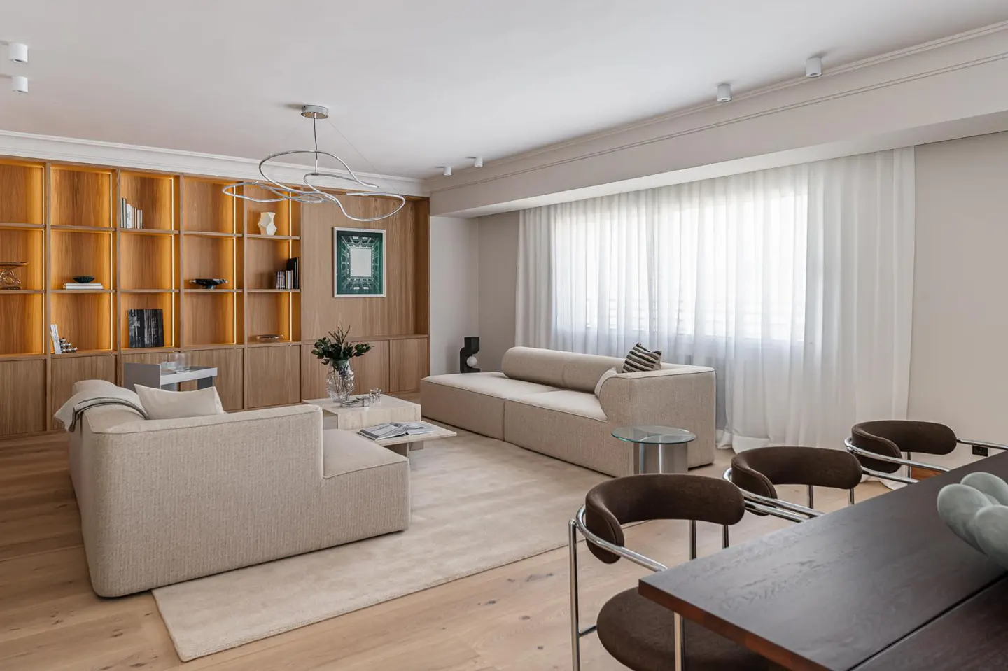 Bright living room with beige sofas, wood floors, and a large built-in bookshelf with warm lighting. Sheer white curtains filter sunlight.