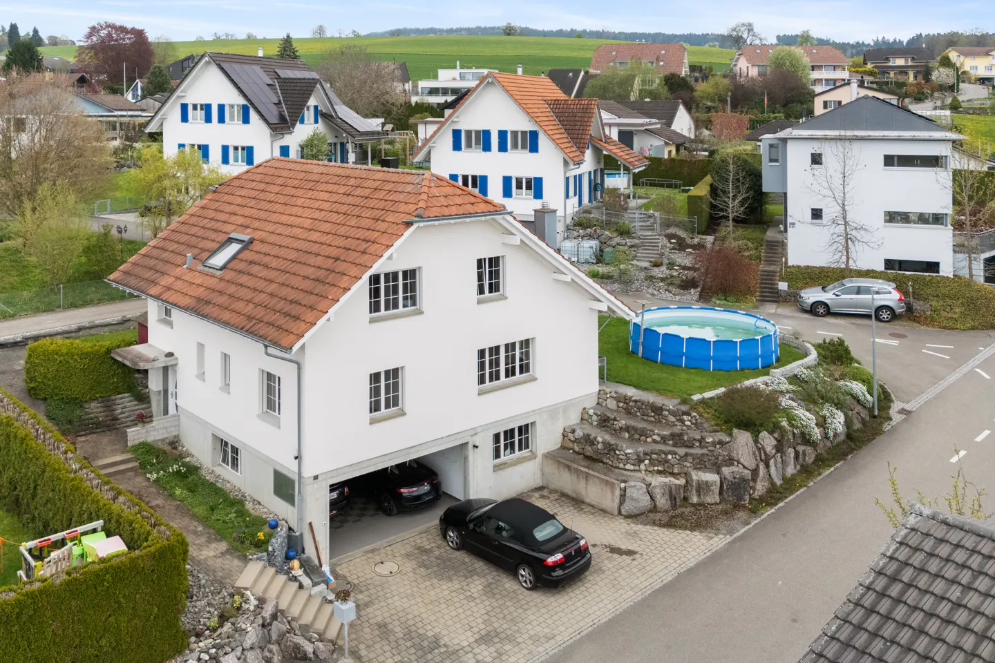 Aerial view of a white two-story house with a red tile roof and an open garage with two cars parked inside. A blue above-ground pool is in the backyard.