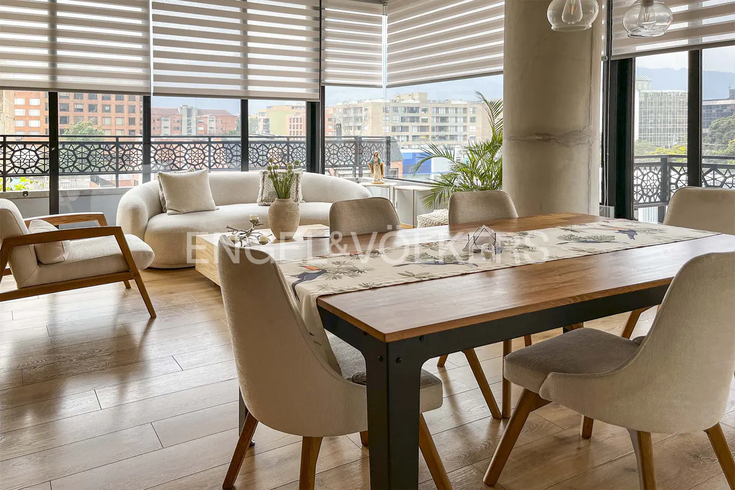Living room with wood floors, a cream sofa, and a wood dining table with beige chairs. Large windows with blinds overlook a city.