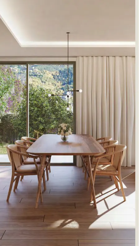 Bright dining room with a wood table, chairs, and a modern chandelier. Large windows overlook a green landscape. Cream-colored curtains add warmth.