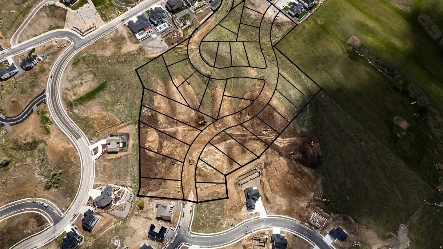Aerial view of a new housing development with dirt lots outlined in black, surrounded by existing homes and green fields. Construction vehicles are visible.