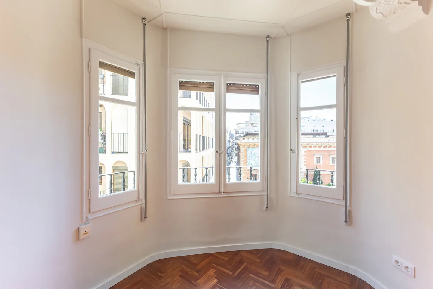 Bright, empty room with herringbone wood floors, white walls, and three white-framed windows showing city views.