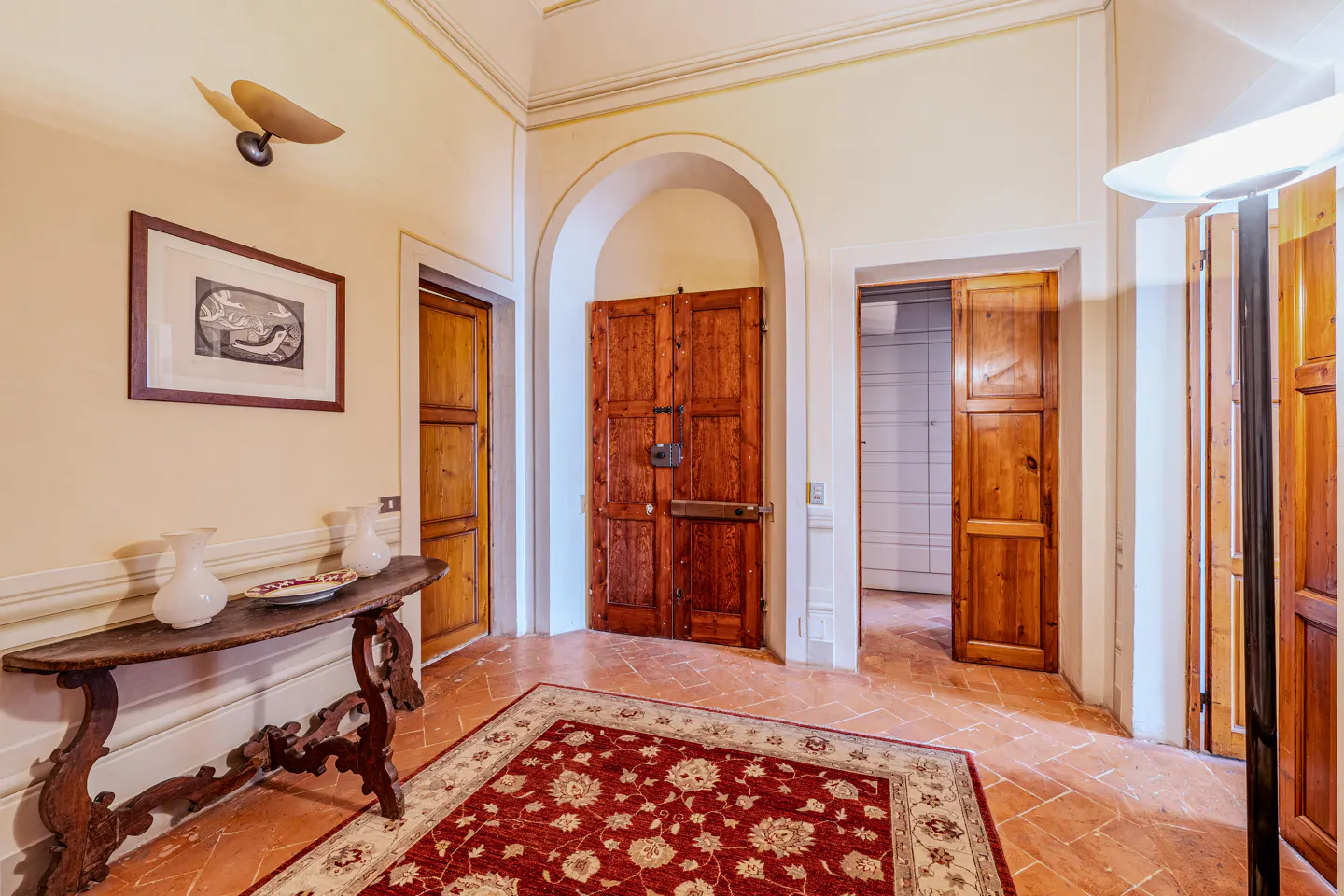 Entryway with terracotta floors, a red patterned rug, wooden doors, and a rustic console table with white vases.