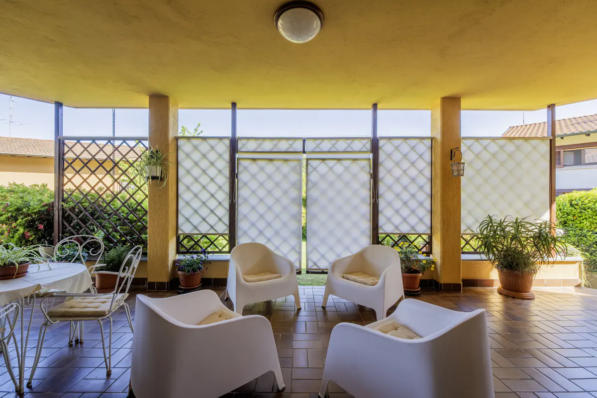 Covered patio with four white chairs, a table and chairs, and potted plants. The patio has a yellow ceiling and a brown tiled floor. A lattice fence surrounds the patio.