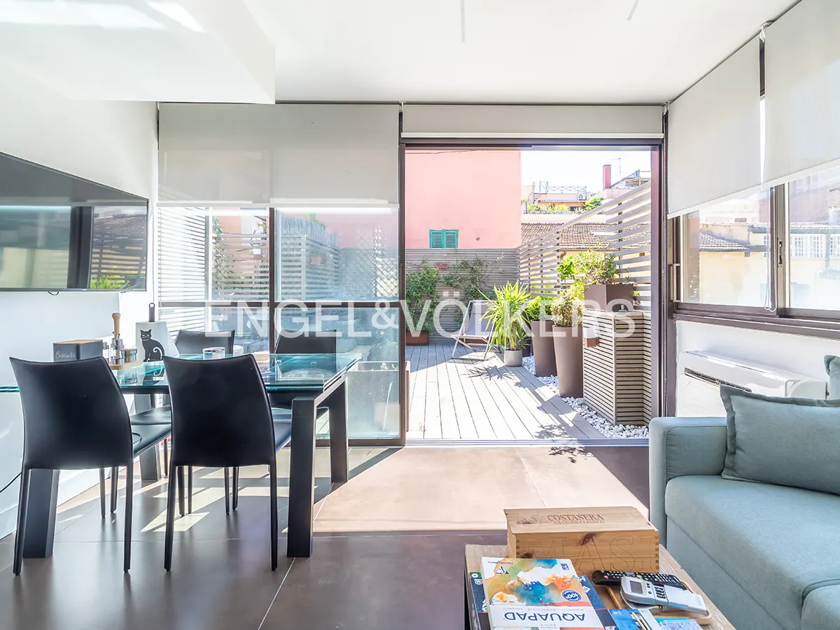 Bright apartment interior with a glass table, black chairs, and a view of a sunny terrace with plants and seating.