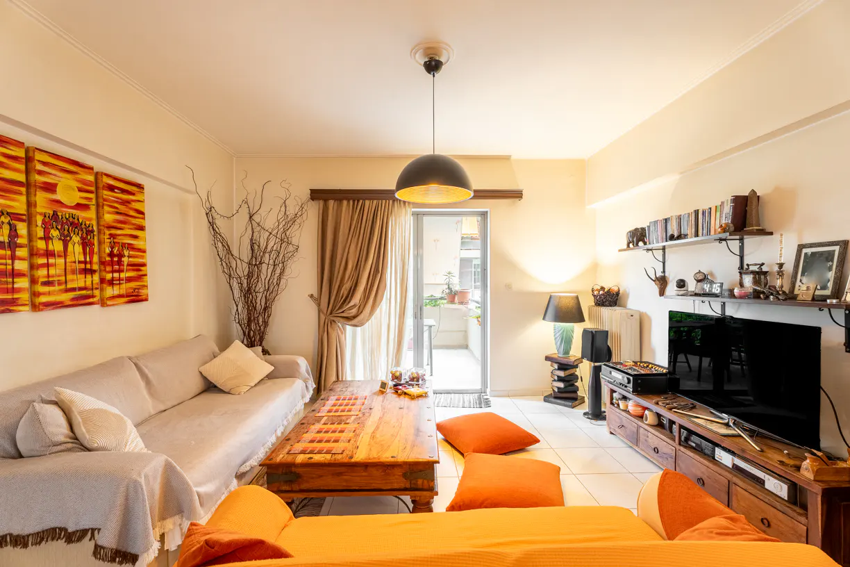 Living room with beige walls, a tan sofa, orange cushions, and a wooden coffee table. A black pendant light hangs from the ceiling. Artwork adorns the walls.
