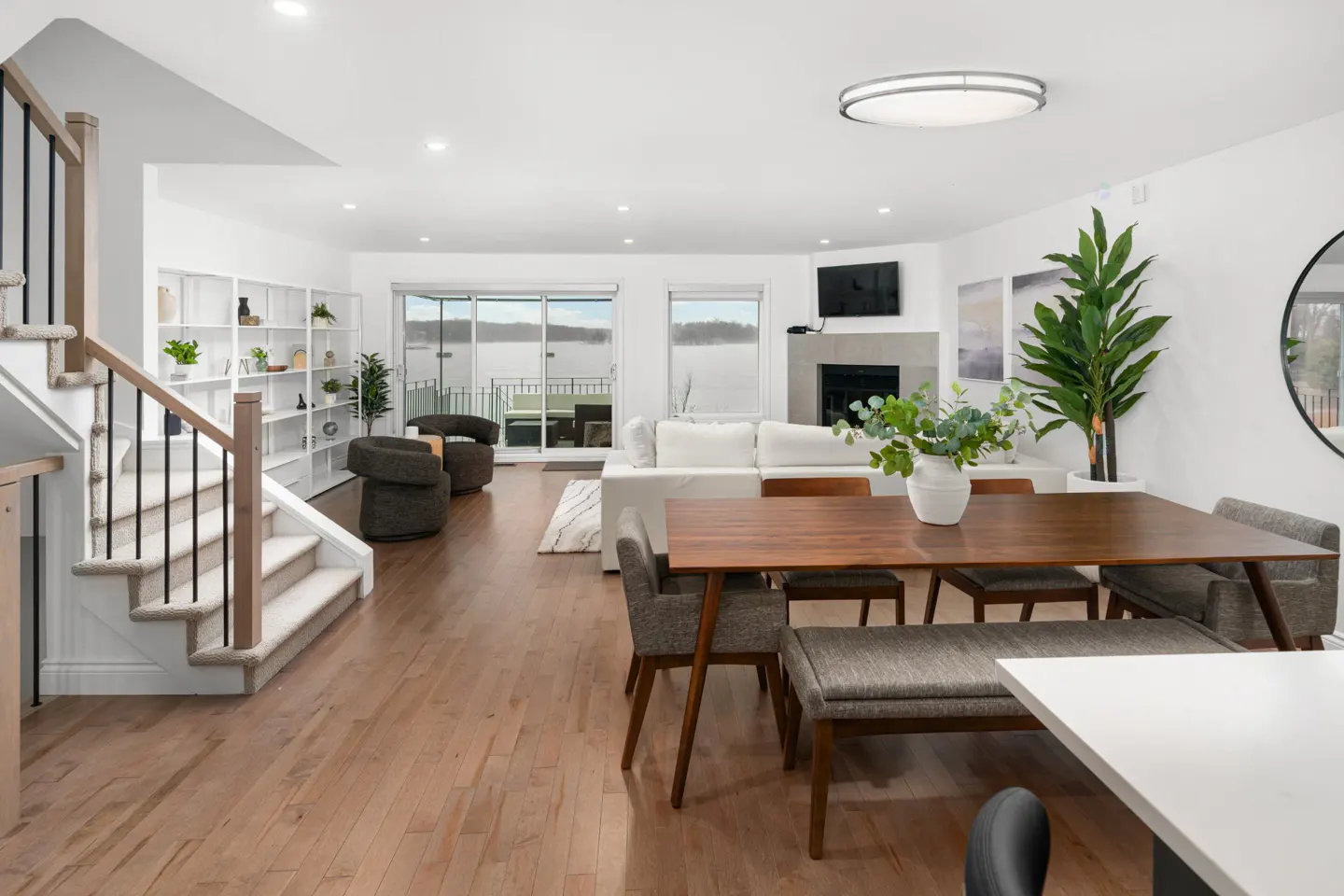 Bright, open-concept living and dining area with hardwood floors, white walls, and large windows overlooking a lake. A wooden dining table sits in the foreground.