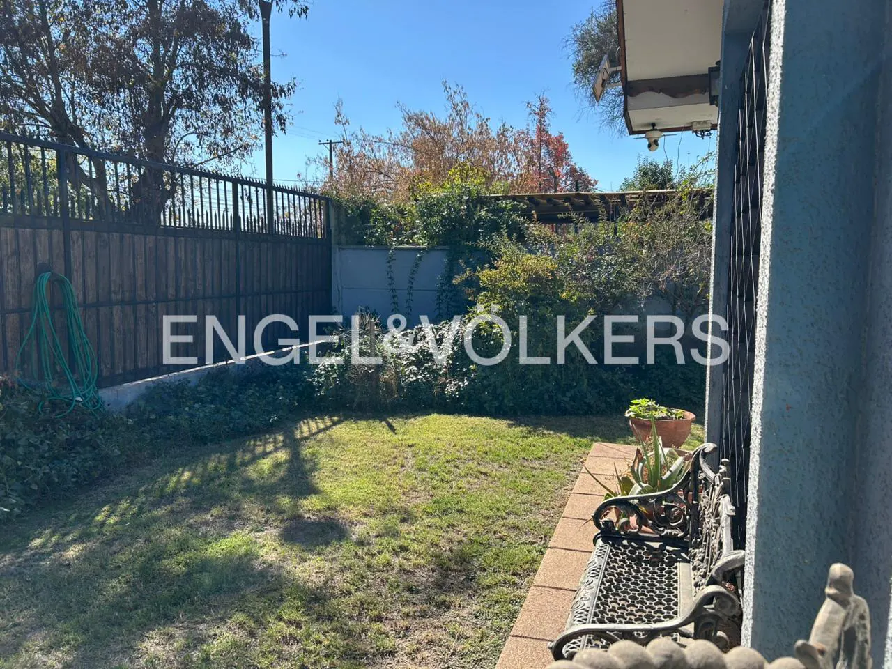A backyard with a metal fence, green grass, and a bench. The Engel & Volkers logo is superimposed on the image.
