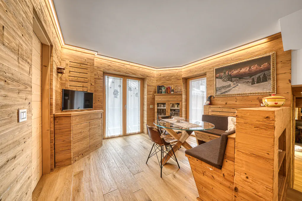 Rustic dining area with wood-paneled walls, a glass-topped table, and built-in seating. A TV and landscape painting add to the cozy ambiance.