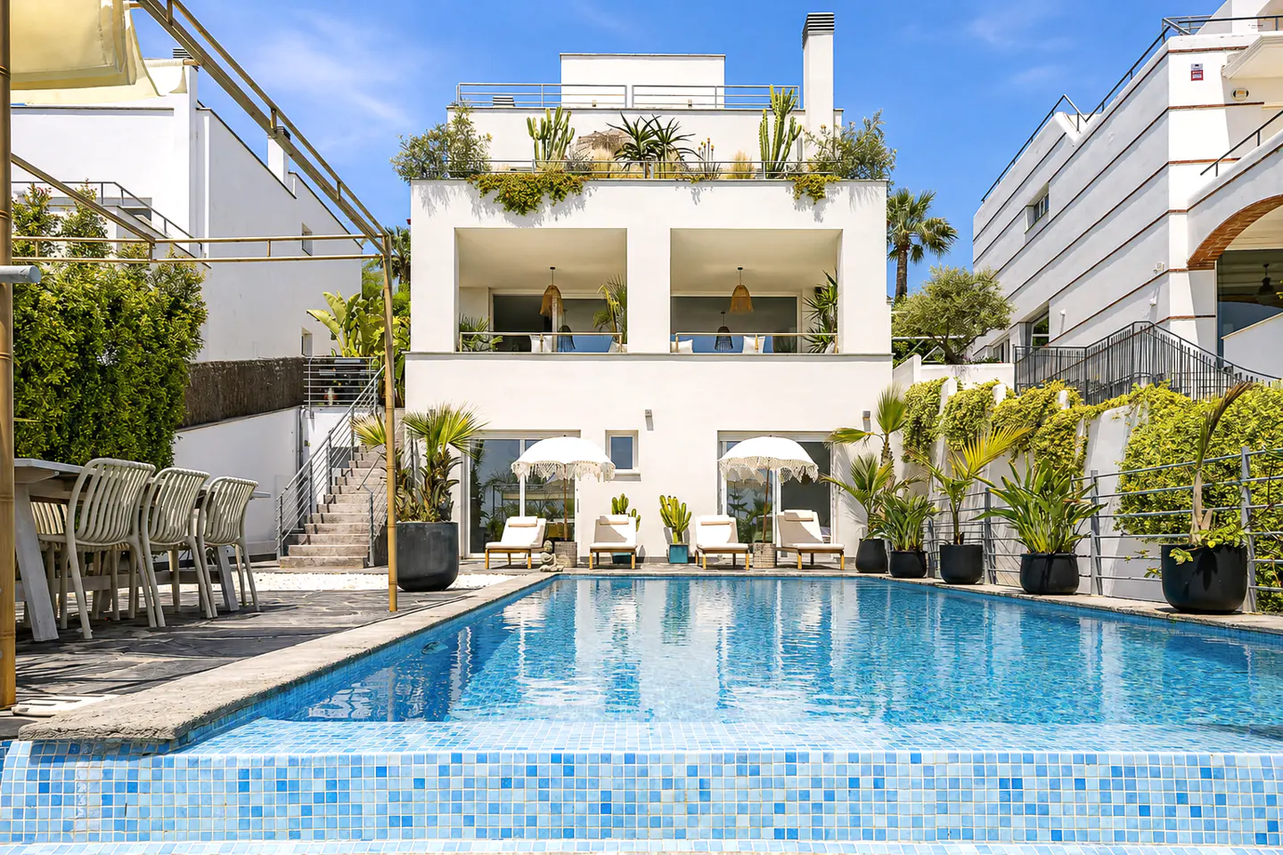 A modern white house with a blue tiled pool, lounge chairs, and plants on the roof.