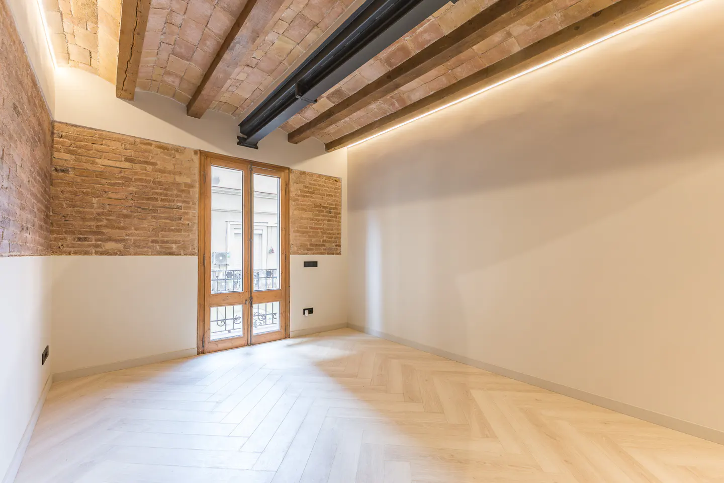 Empty room with herringbone wood floors, brick and plaster walls, and exposed wood beam ceiling. Double doors lead to a balcony.