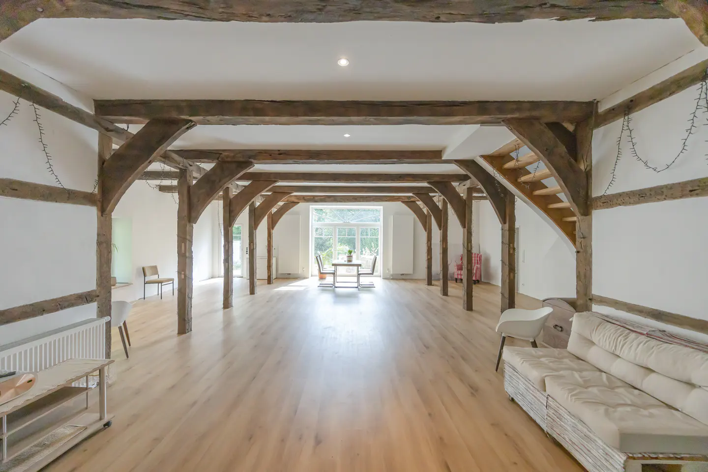 Bright, open room with wooden beams, light wood floors, and white walls. A table and chairs are visible in the distance. A white couch is in the foreground.