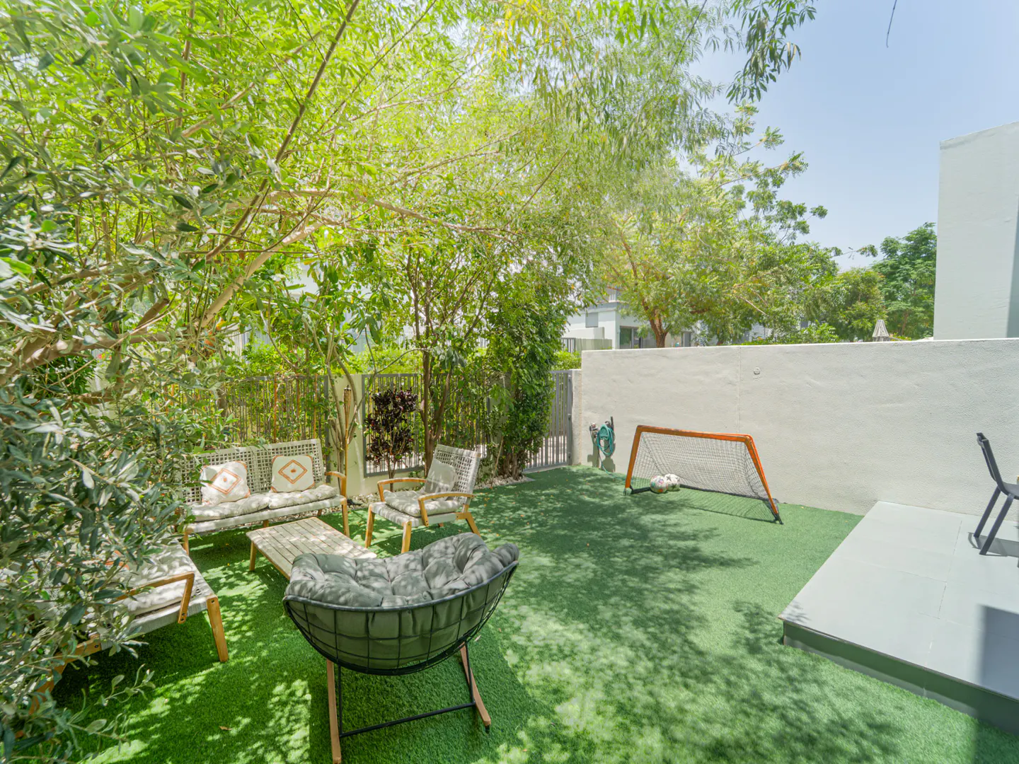 A sunny backyard with artificial turf, patio furniture, a small soccer goal, and lush trees against a white wall.