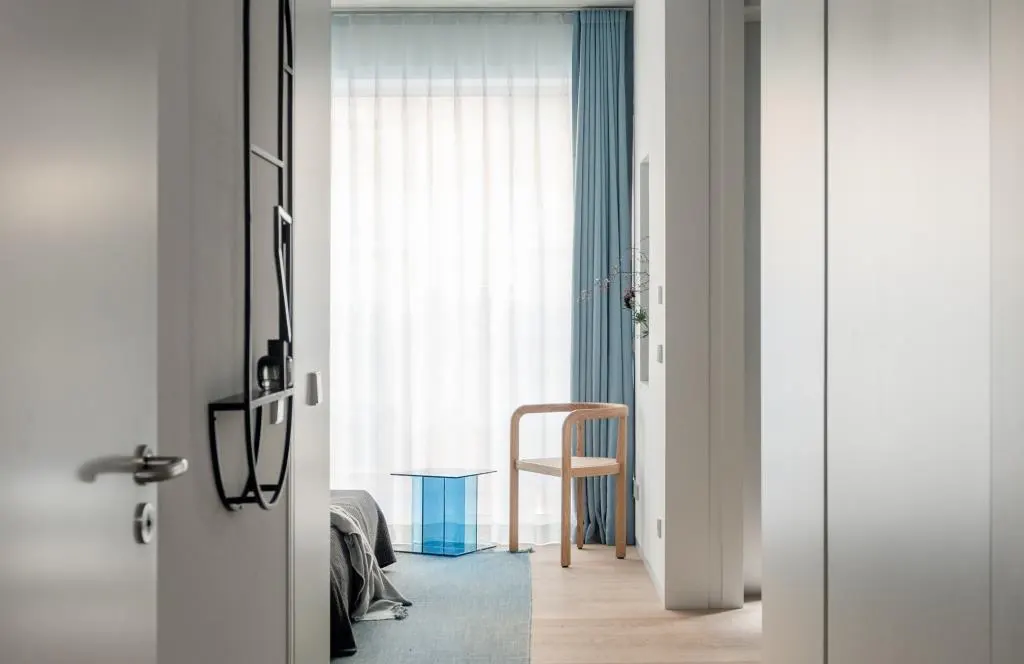 A view into a modern bedroom with a blue glass table, a wooden chair, and white curtains. A black metal shelf is on the wall.