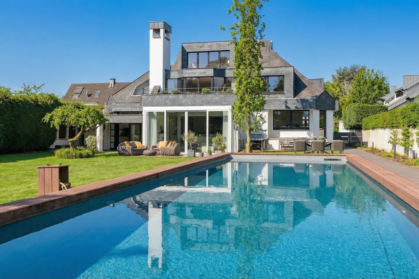 A modern two-story house with a pool. The house is white with a gray roof and a tall white chimney. The pool is blue and reflects the house.