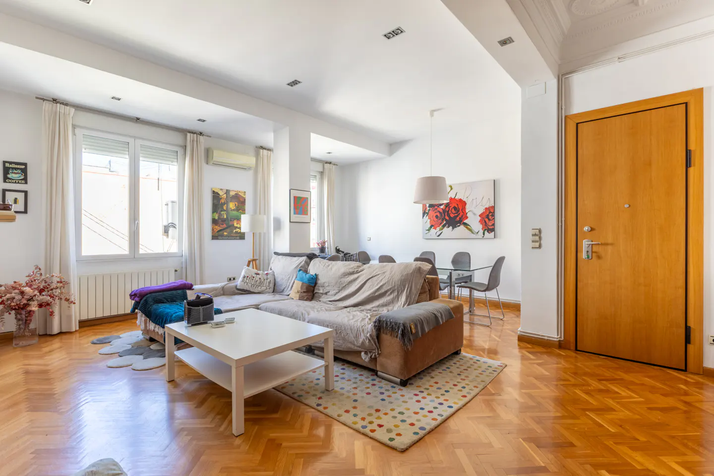 Bright living room with wood floors, white walls, and a large sectional sofa. A white coffee table sits on a colorful rug. Dining table and art are visible.