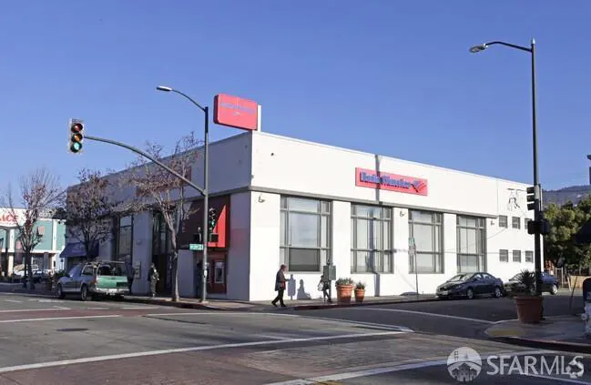 Street view of a Bank of America building with a white facade and large windows. A man walks across the crosswalk. Cars are parked nearby.