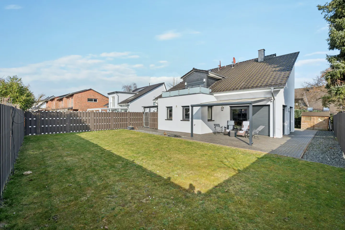 A white house with a gray roof overlooks a green lawn, enclosed by a brown fence. A patio with chairs sits outside the house.