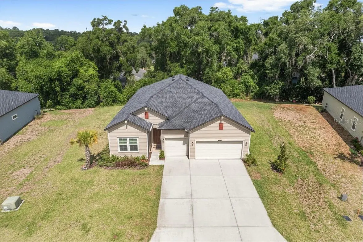 Aerial view of a beige single-family home with a gray roof, a two-car garage, and a concrete driveway, surrounded by a green lawn and trees.
