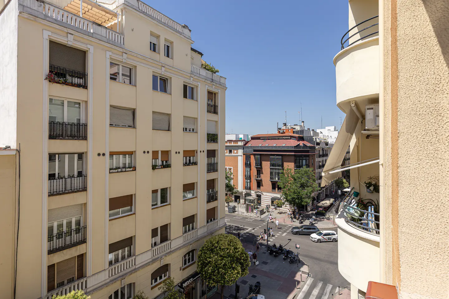 View from a balcony overlooking a sunny city street intersection with cream-colored apartment buildings and traffic.