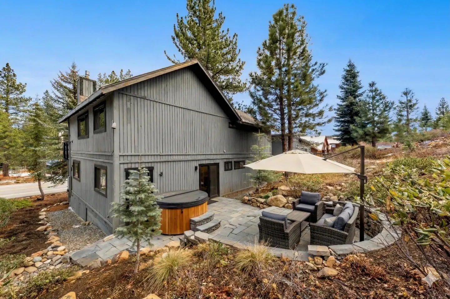 Two-story gray house with a hot tub and patio set under a white umbrella. Tall pine trees surround the property.