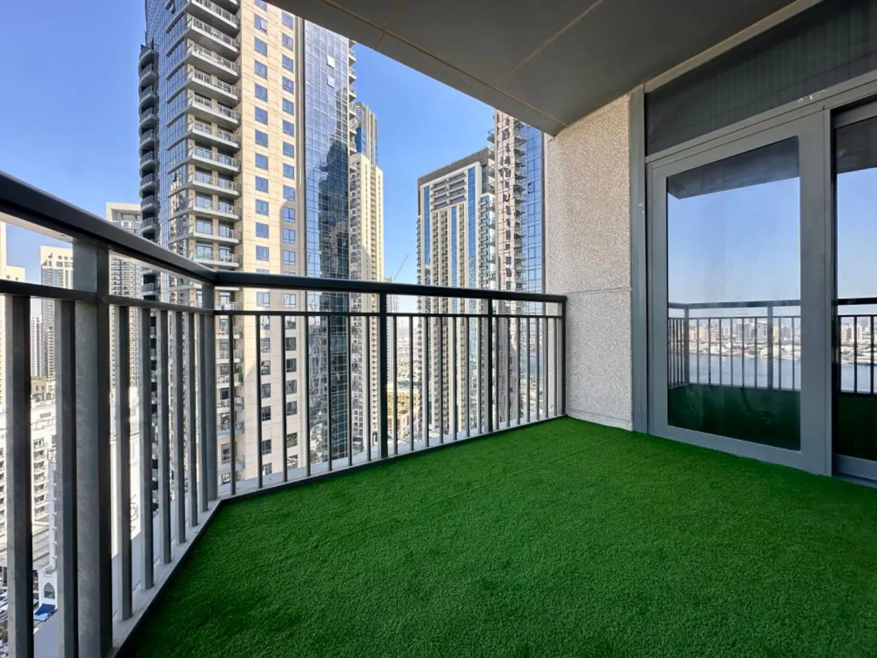 Balcony with green turf, metal railing, and sliding glass doors. City skyline visible in the background.