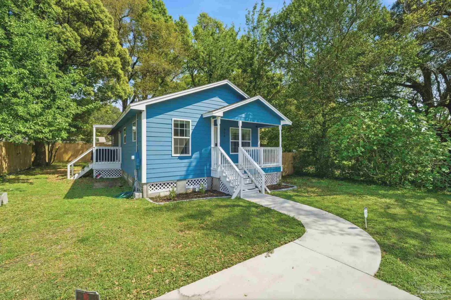 A blue, one-story house with white trim and a front porch is surrounded by green grass and trees. A curved, concrete walkway leads to the house.