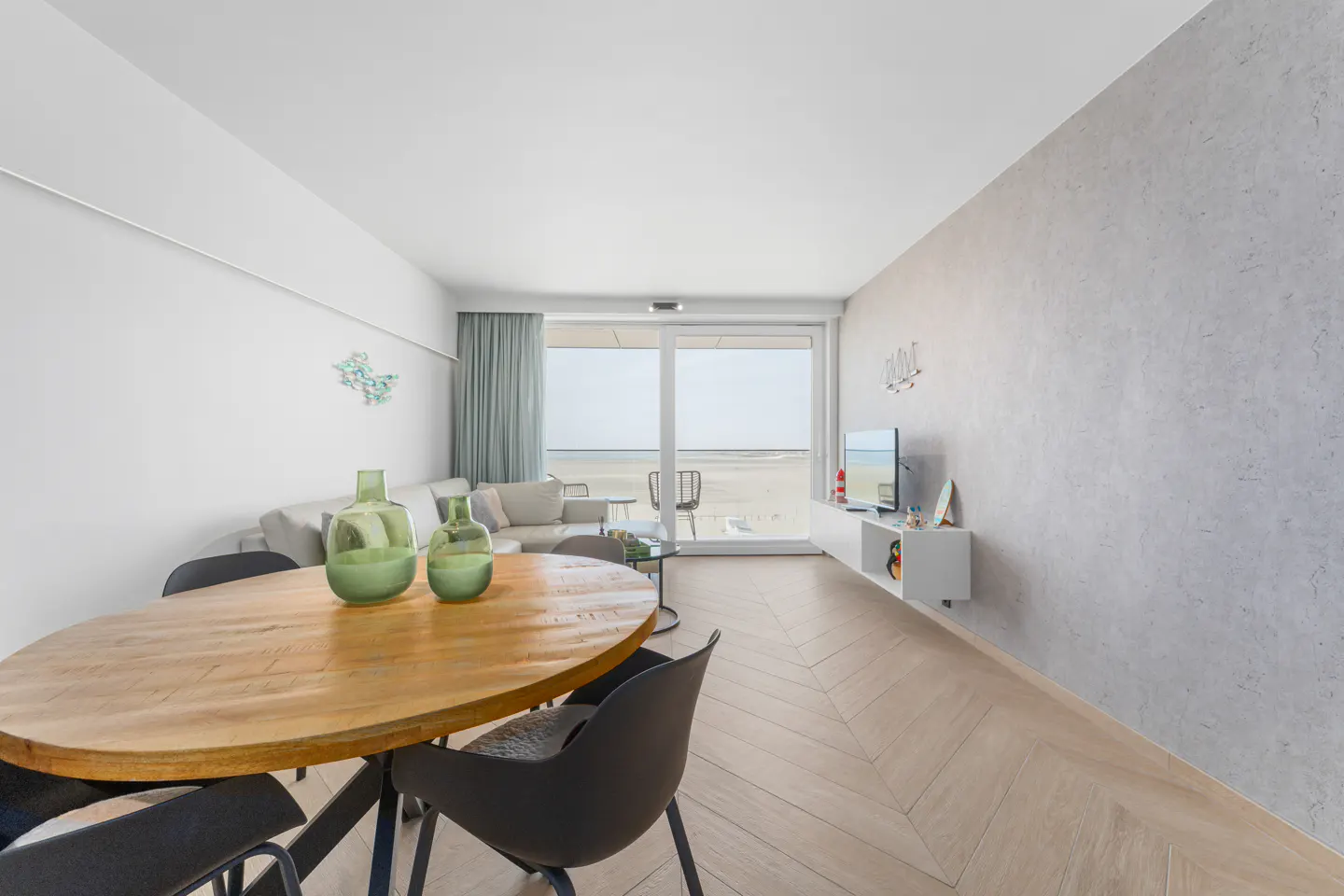 Bright living room with a wooden table, black chairs, and green vases. A sofa and TV are visible, with a beach view through a sliding glass door.