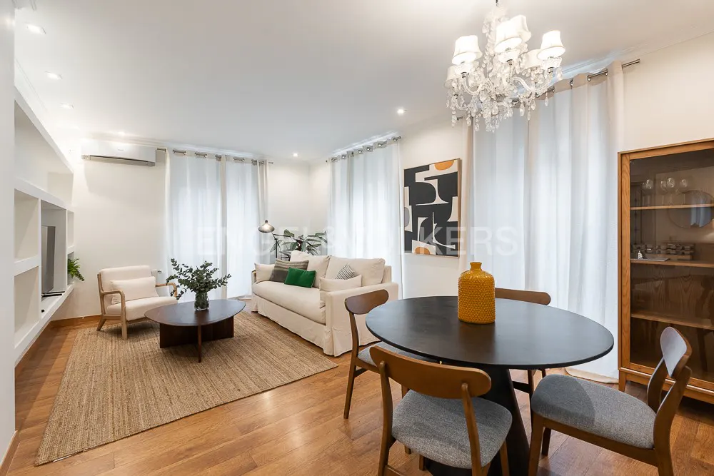 Bright living room with hardwood floors, white walls, and sheer curtains. A sofa, chairs, and a round dining table furnish the space. A crystal chandelier hangs above.