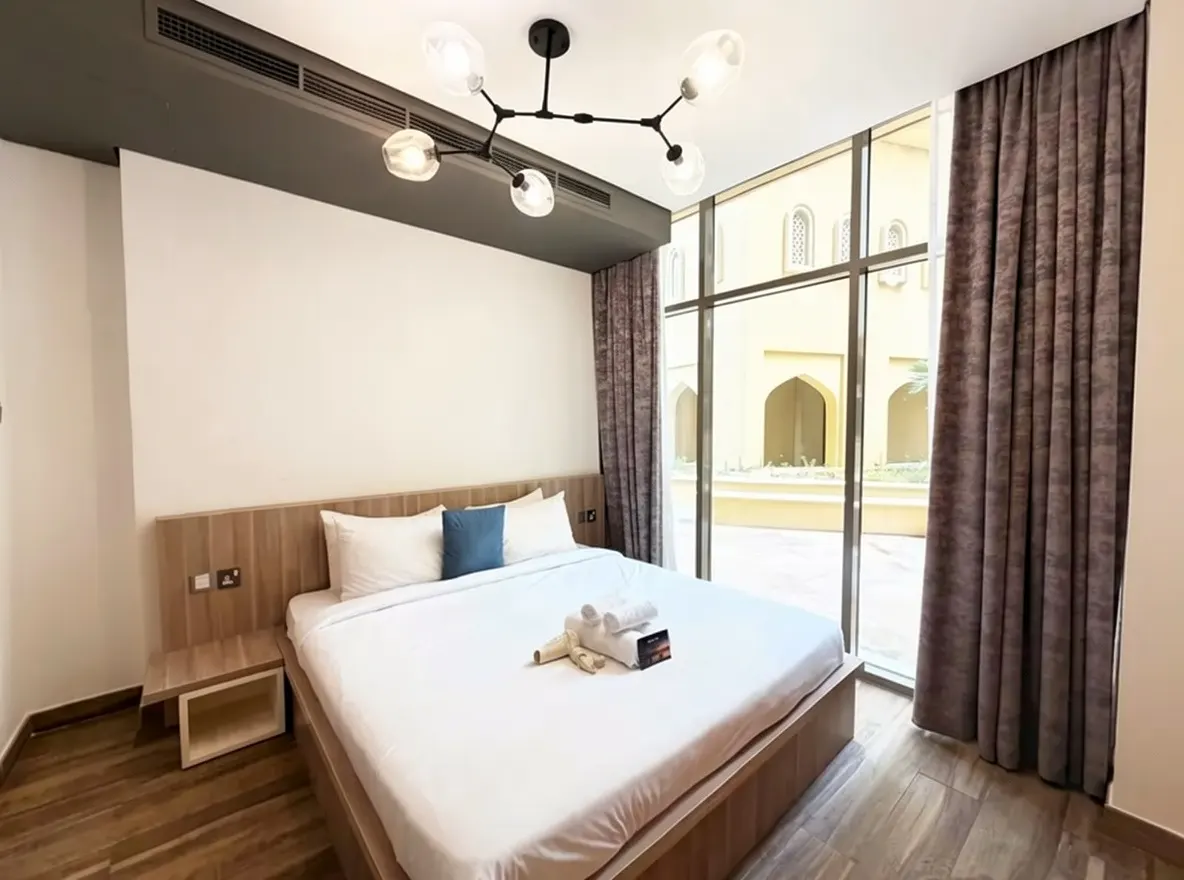 A bedroom with a white bed, blue pillow, and towels. A large window shows an exterior courtyard.