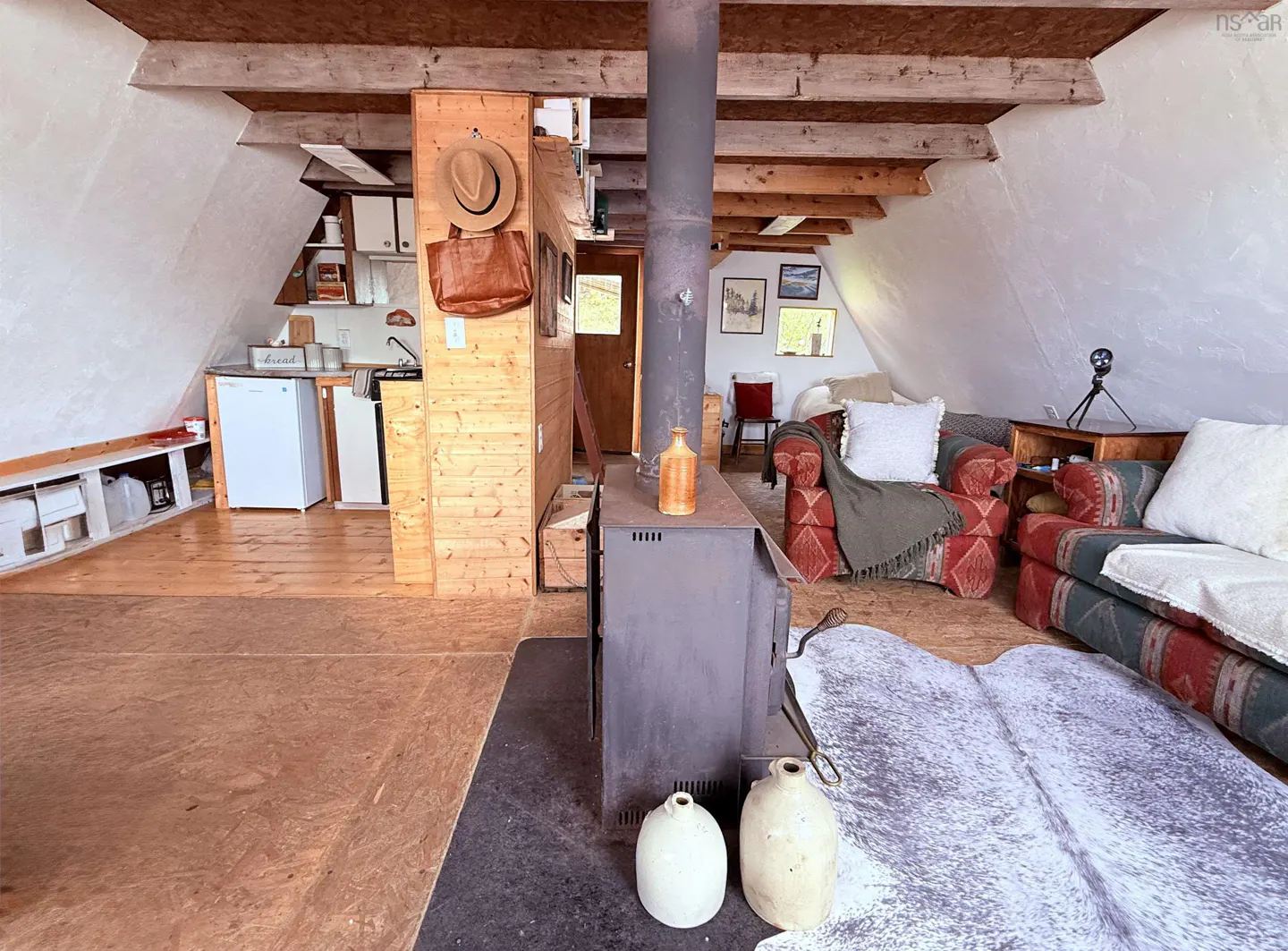 A-frame cabin interior with wood stove, kitchen, and seating area. A hat and bag hang on the wood wall.