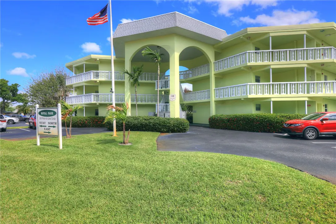 Exterior of a light green, three-story condo building with white balconies and an American flag. A sign reads "Royal Park." A red car is parked nearby.