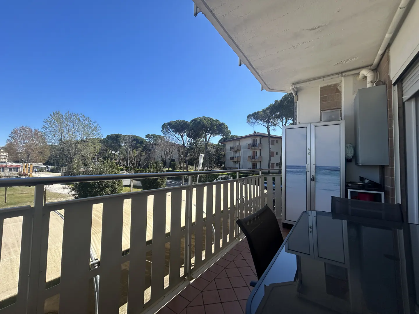 Balcony view with a black table, chairs, and a metal railing overlooking trees and buildings under a clear blue sky.