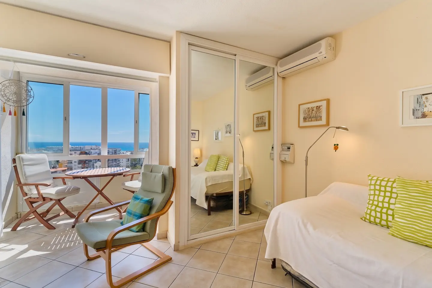 Bright apartment interior with a balcony overlooking the sea. A mirrored closet reflects a bed. A daybed with green pillows sits nearby.