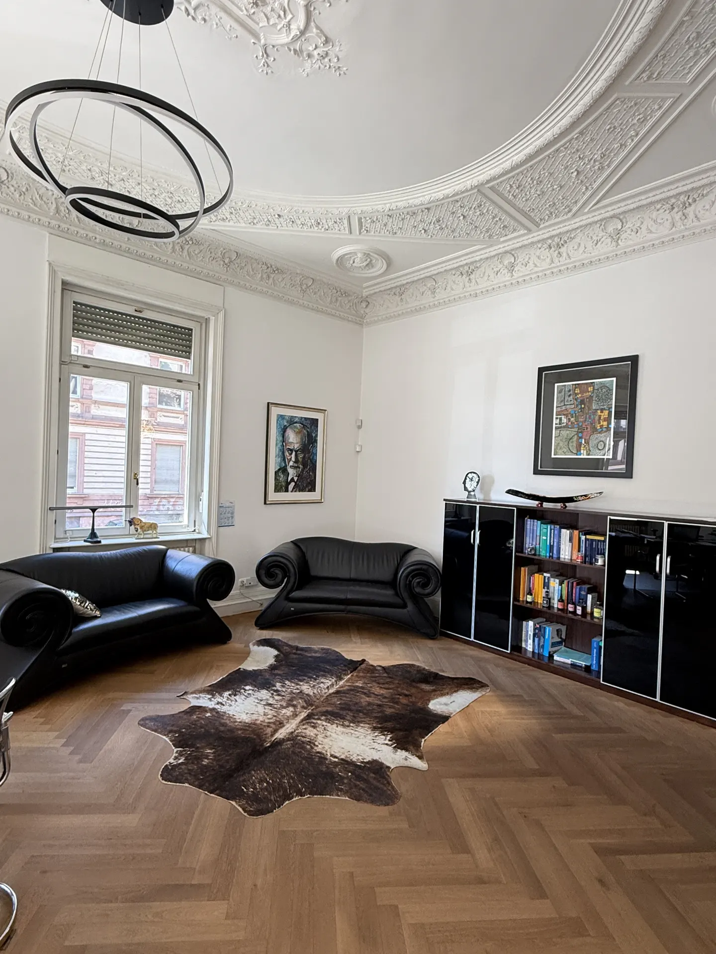 Living room with two black sofas, a cowhide rug, and a black cabinet with books. A modern chandelier hangs from the ornate ceiling.