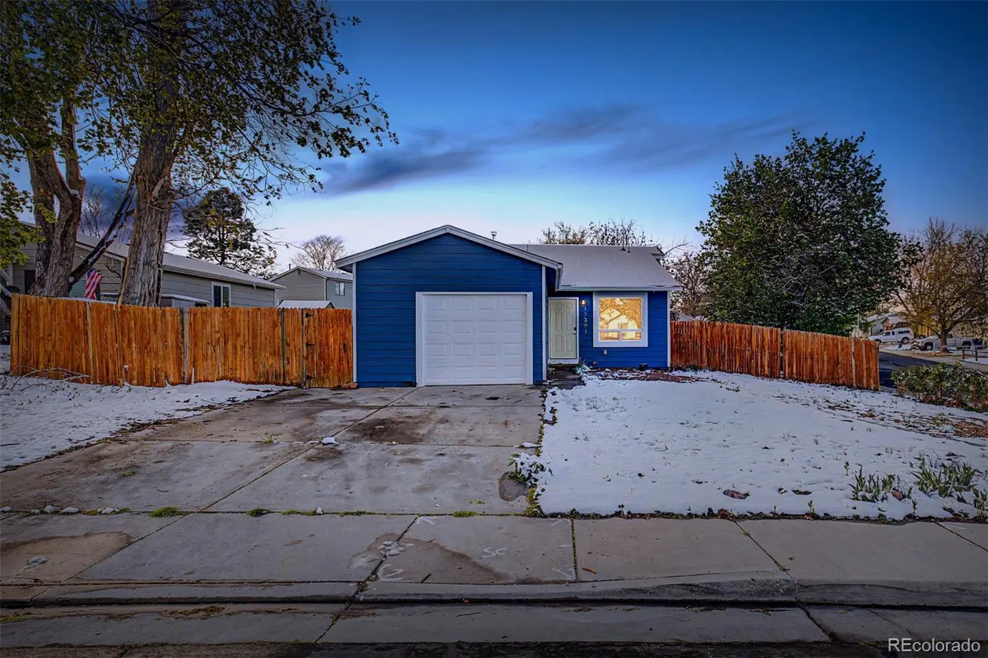 A blue single-story house with a white garage door and a wooden fence, with snow on the ground.