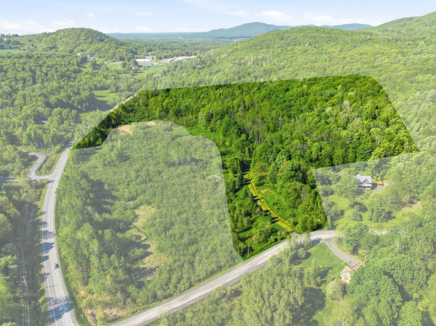 Aerial view of a large, wooded plot of land highlighted in green, situated near a road and rolling green mountains.