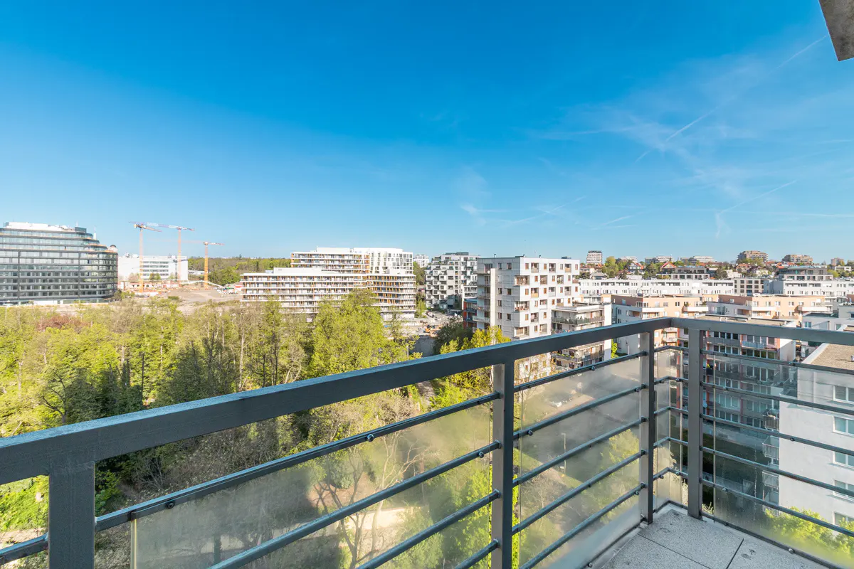 View from a balcony with a gray metal railing overlooking a city with white buildings and green trees under a clear blue sky.