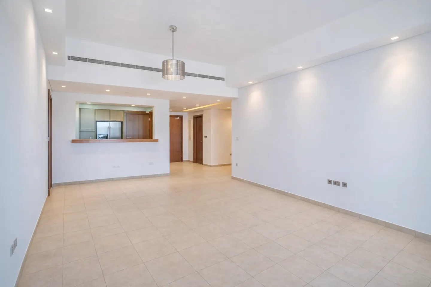 Bright, empty living room with white walls, beige tile floor, and a view into the kitchen with wood cabinets.