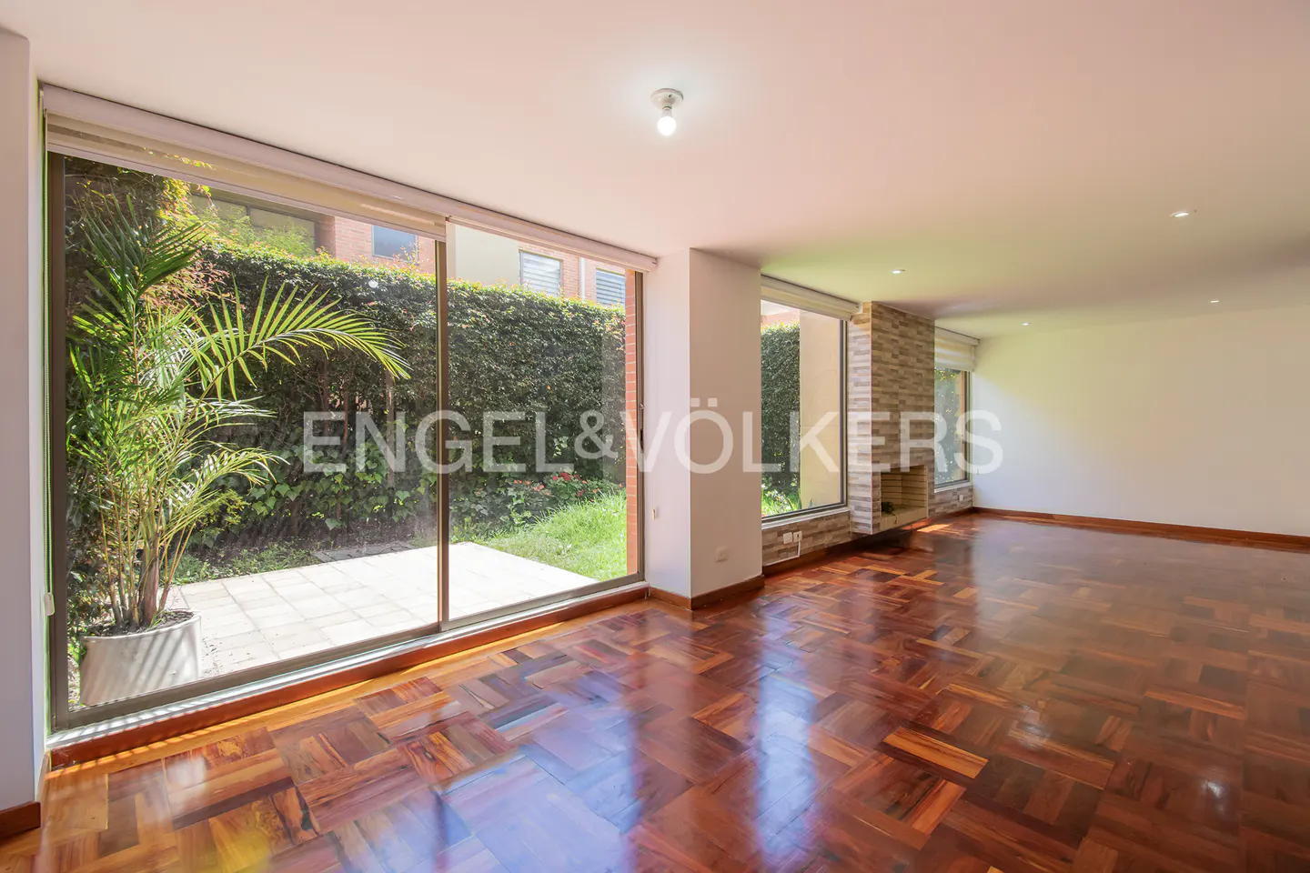 An empty living room with hardwood floors, a large window overlooking a green garden, and a stone fireplace.