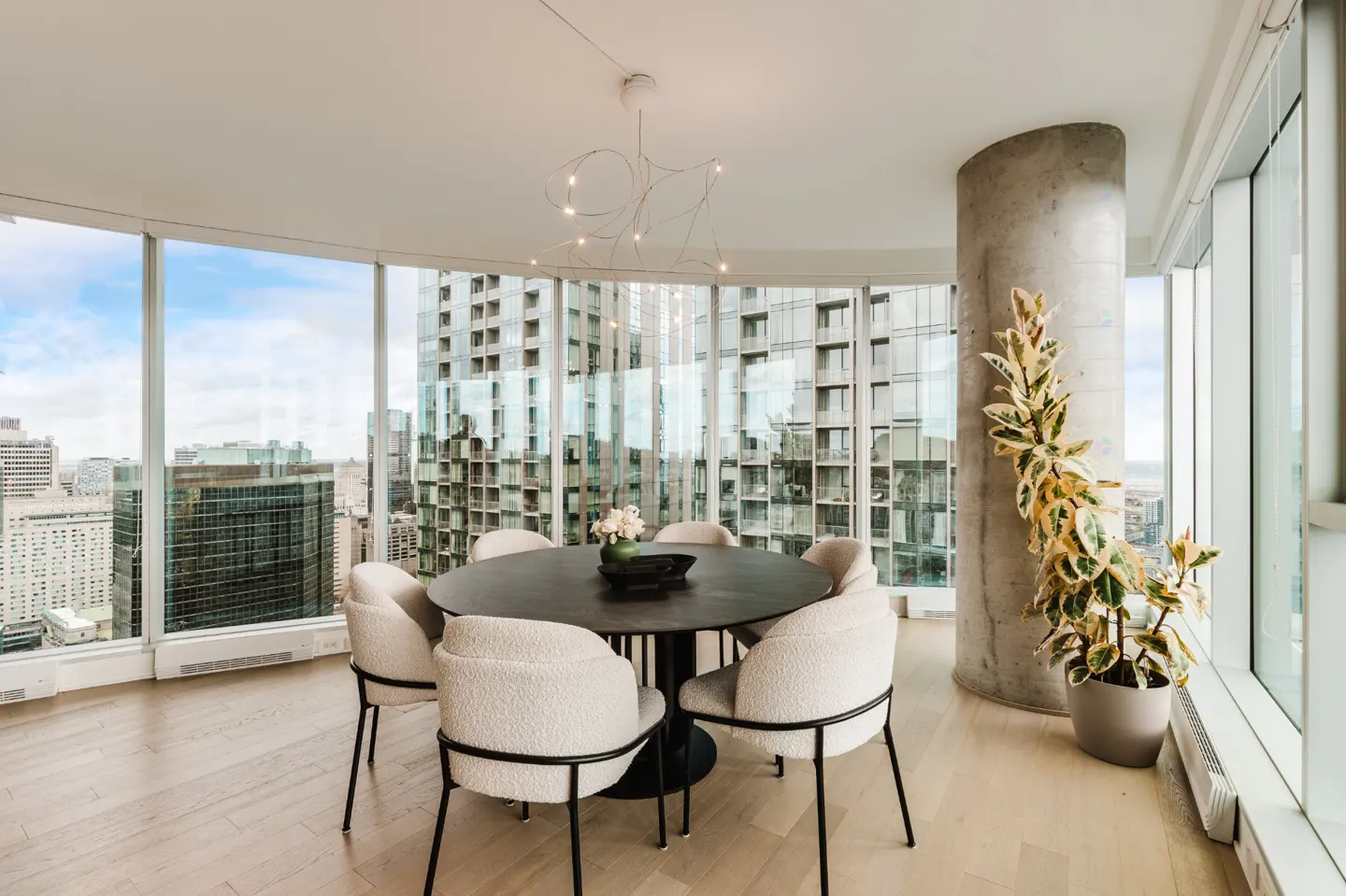Bright dining room with city view. Round table with white chairs, modern light fixture, and potted plant by a concrete pillar.