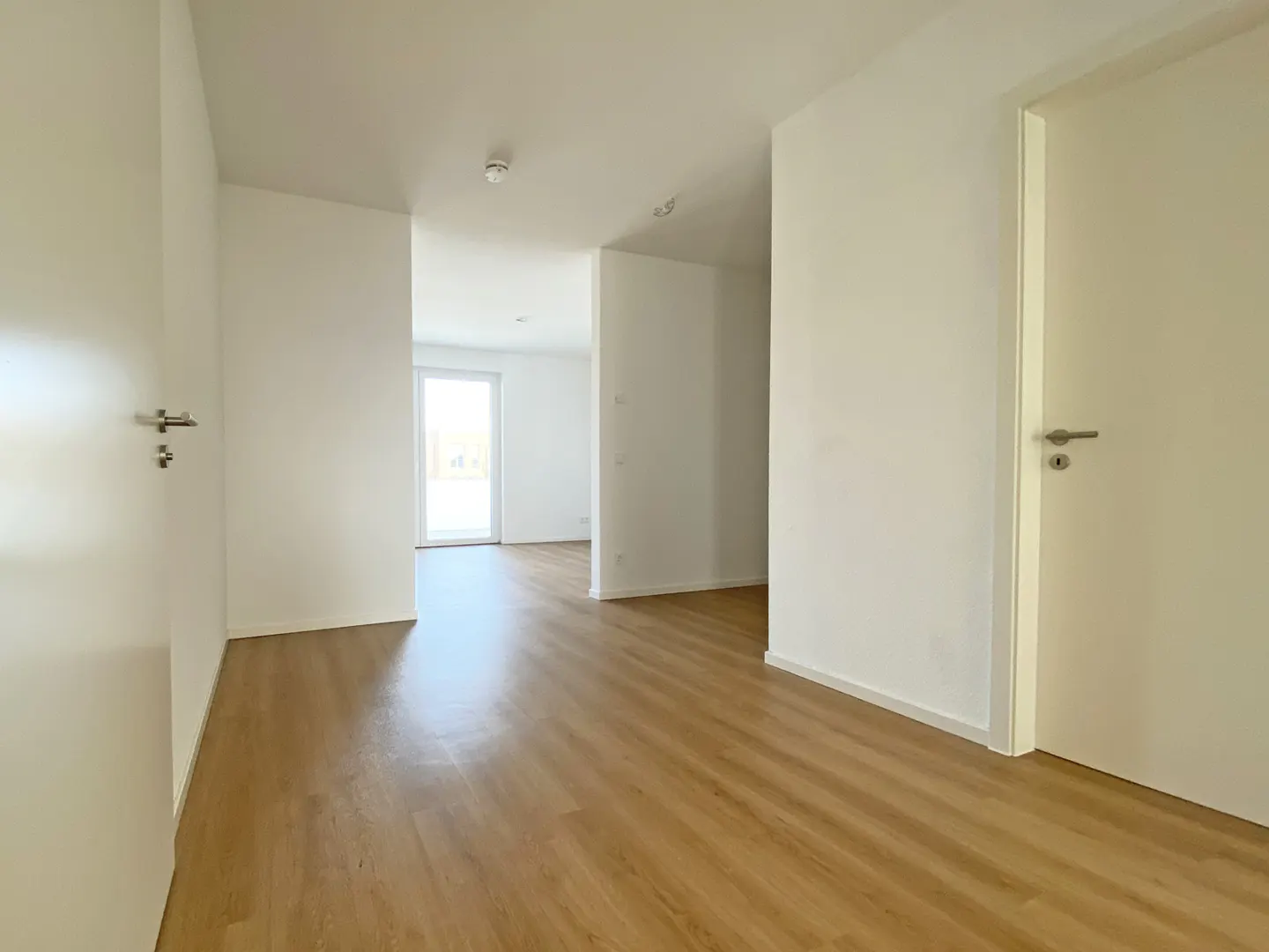 Bright, empty hallway with light wood floors and white walls. A door is on the right, and a window is visible at the end of the hall.