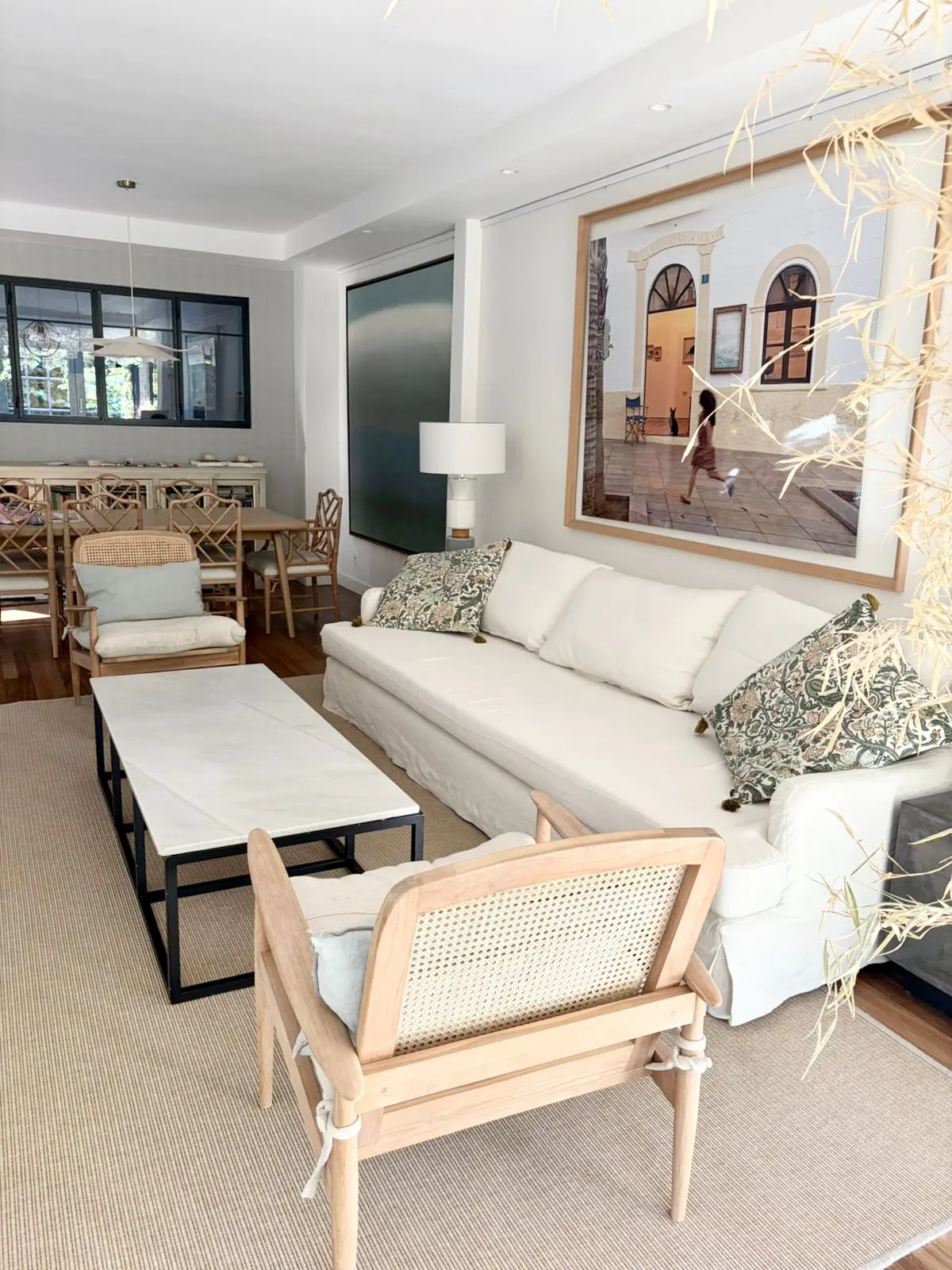 Bright living room with a white sofa, light wood chairs, and a marble coffee table on a beige rug. A large framed photo hangs above the sofa.