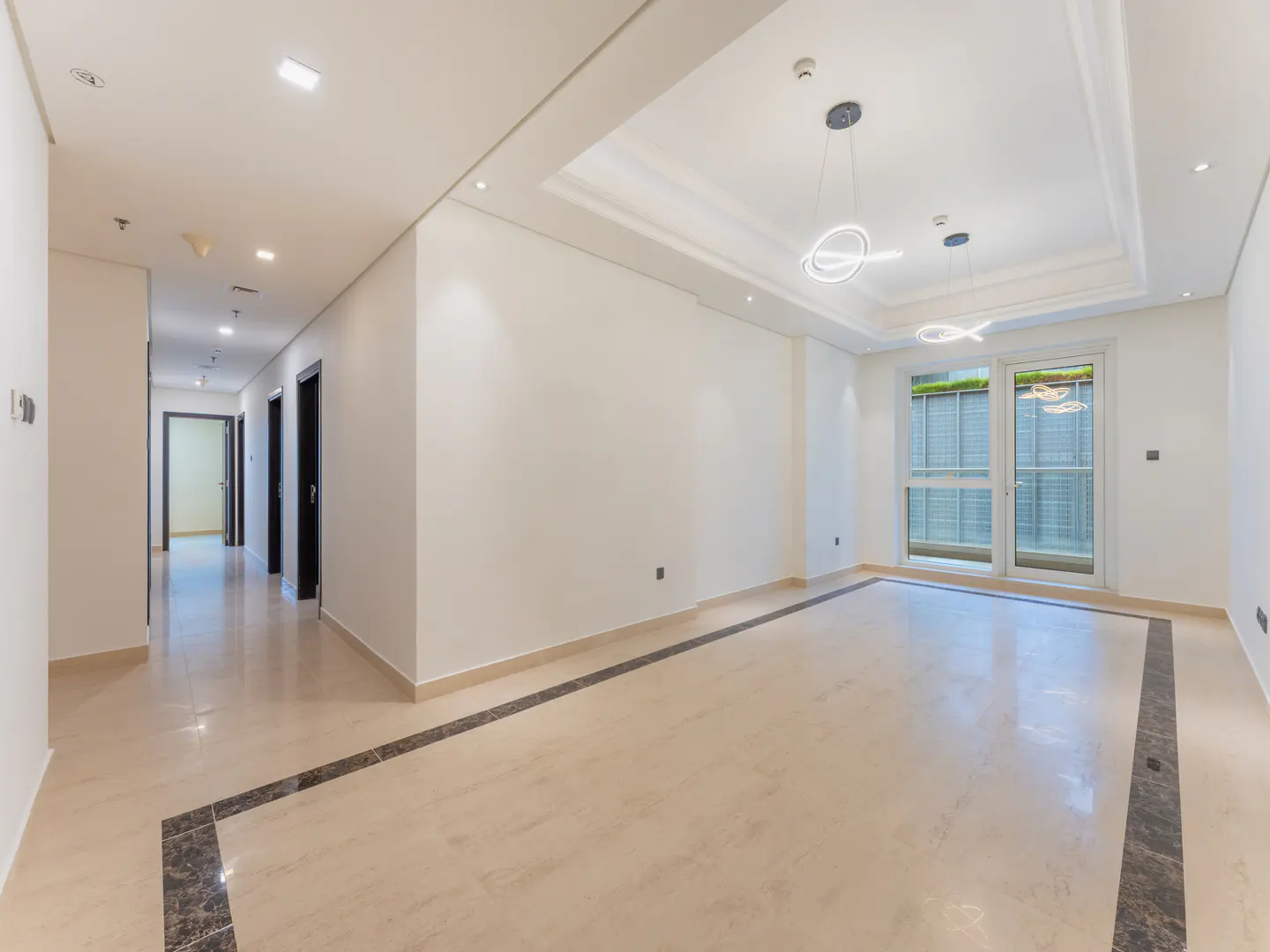 Bright, empty room with marble floors and white walls. A hallway with dark doors is visible on the left. Modern light fixtures hang from the ceiling.