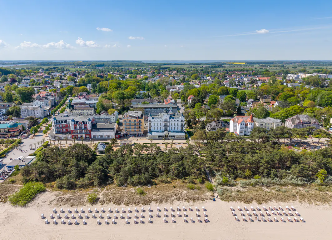 Aerial view of a beach with rows of white chairs, a line of trees, and buildings in the background.