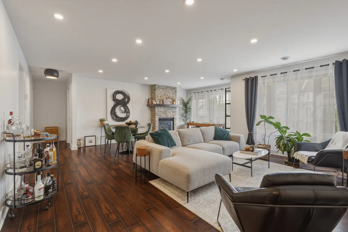 Bright living room with hardwood floors, a beige sectional sofa, stone fireplace, and a bar cart with bottles.