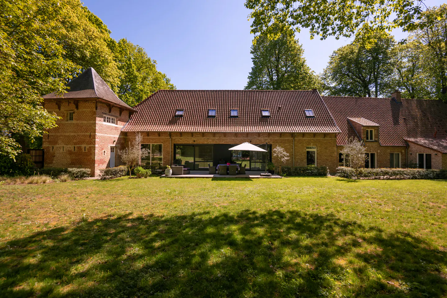 Exterior view of a brick house with a red tile roof, a turret, and a green lawn. A patio with furniture is visible.