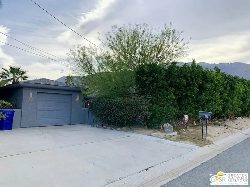A gray house with a garage and a concrete driveway, surrounded by green bushes and trees. Mountains are visible in the background.